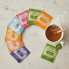 Colorful Yogi tea bags from the wellness sampler fanned out on a white marble surface next to a steaming white ceramic mug of herbal tea.