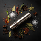Prima Cucina Battery-Operated Pepper Mill flat lay on dark slate surface surrounded by whole black peppercorns, coarse sea salt, and dried spices, overhead moody kitchen shot