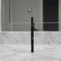 Mary Kay Liquid Eyeliner Pen in MK Black — sleek glossy black barrel with silver Mary Kay lettering standing upright on a white marble vanity with soft dramatic lighting and blurred mirror
