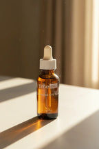 Small dark amber glass skincare dropper bottle sitting in bright warm natural sunlight on smooth stone surface