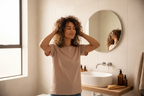 Woman with wavy coily hair scrunching styling cream into damp hair in bright modern bathroom curl routine lifestyle photo
