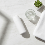 Lifestyle — white electric facial cleansing brush on white marble vanity beside glass of water, white towel, and small succulent — clean minimalist skincare editorial for visual context only