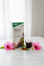 Salus Floradix Floravital Liquid Iron and Vitamin Formula 500ml retail box on a marble surface with hibiscus flowers, chamomile, rose hips, and a small glass of dark liquid supplement – More4less Outlet
