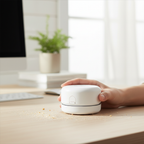 Person's hand holding a compact white circular mini desktop vacuum over a wooden desk surface with crumbs and dust