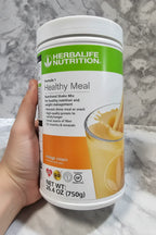 In-hand close-up of Herbalife Formula 1 Orange Cream canister on white marble surface