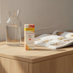 Burt's Bees Truly Glowing Glow Booster box on minimalist light-oak dressing table with clear glass carafe of water and open lifestyle magazine