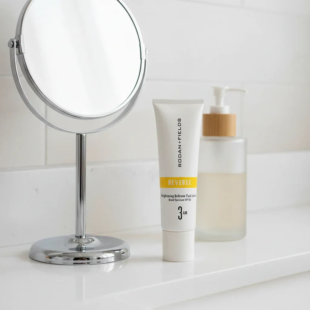 Rodan + Fields Reverse Brightening Defense Fluid SPF 50 tube on minimalist white quartz bathroom vanity shelf with designer handheld mirror and premium skincare dispenser