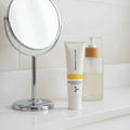 Rodan + Fields Reverse Brightening Defense Fluid SPF 50 tube on minimalist white quartz bathroom vanity shelf with designer handheld mirror and premium skincare dispenser
