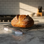 Freshly baked sourdough loaf with open ear crust on wire cooling rack beside grey Breadsmart bread lame on marble countertop