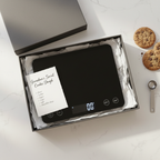 Lifestyle — black digital food scale in gift box with white tissue paper and handwritten recipe card on white kitchen counter — gifting flatlay editorial for visual context only