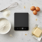 Lifestyle — overhead flatlay of black digital kitchen scale surrounded by eggs, flour bowl, butter, and whisk on white marble — editorial for visual context only