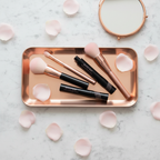 Mary Kay Lash Love Lengthening Mascara I Love Black glossy black tube with pink branding on a rose-gold vanity tray with makeup brushes, a small mirror, and pink rose petals on white marble — feminine beauty flat-lay