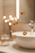 Amber serum droplets suspended mid-air against a soft bokeh light bathroom vanity background in warm soft lighting