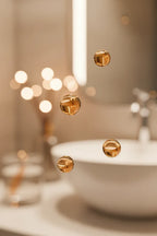 Amber serum droplets suspended mid-air against a soft bokeh light bathroom vanity background in warm soft lighting