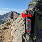 Orange portable air horn signal device clipped to hiking backpack strap on mountain trail, outdoor safety lifestyle