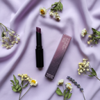 Mary Kay Lip Suede Lipstick Mulberry Muse black tube with cap off next to its original pink-and-grey retail box on soft lavender fabric with scattered dried flowers — feminine lifestyle flat-lay