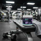 Alani Nu Cosmic Stardust Pre-Workout tub on training gym bench with dumbbells, towel, and water bottle.