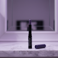 Mary Kay Smoke & Shimmer Eye Wand in Amethyst Smoke — glossy black barrel with dark purple cap beside it, metallic slate-purple cream tip twisted up, standing upright on a white marble vanity with soft moody purple-toned lighting and blurred mirror