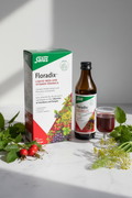Salus Floradix Iron and Herbs Liquid Formula 500ml retail box on a marble surface with rose hips, stinging nettle, fennel, and a small glass of dark red liquid supplement – More4less Outlet