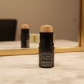 Mary Kay Glowing Finish Illuminating Stick in Gold — glossy black cylindrical stick with cream golden-bronze shimmer bullet twisted up, standing upright on a white marble vanity with warm golden lighting and blurred mirror