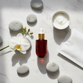 Lifestyle image - elegant red glass serum bottle beside white orchid and river stones on marble, Japanese skincare editorial flatlay for visual context only