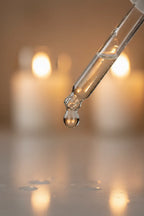 Detail shot of a clean glass dropper with clear liquid serum drop reflecting warm indoor lifestyle lighting with soft bokeh background