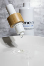 Dispensing pump releasing a clear watery hyaluronic acid serum drop onto smooth white porcelain in soft clean lighting