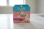 Gillette Venus Comfortglide White Tea 4-count refill box on white marble bathroom surface — light blue and pink packaging with purple Comfortglide banner and seashell illustration