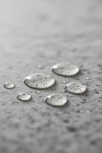 Clear droplets of a professional facial hydration serum on polished grey stone surface in soft diffused lighting