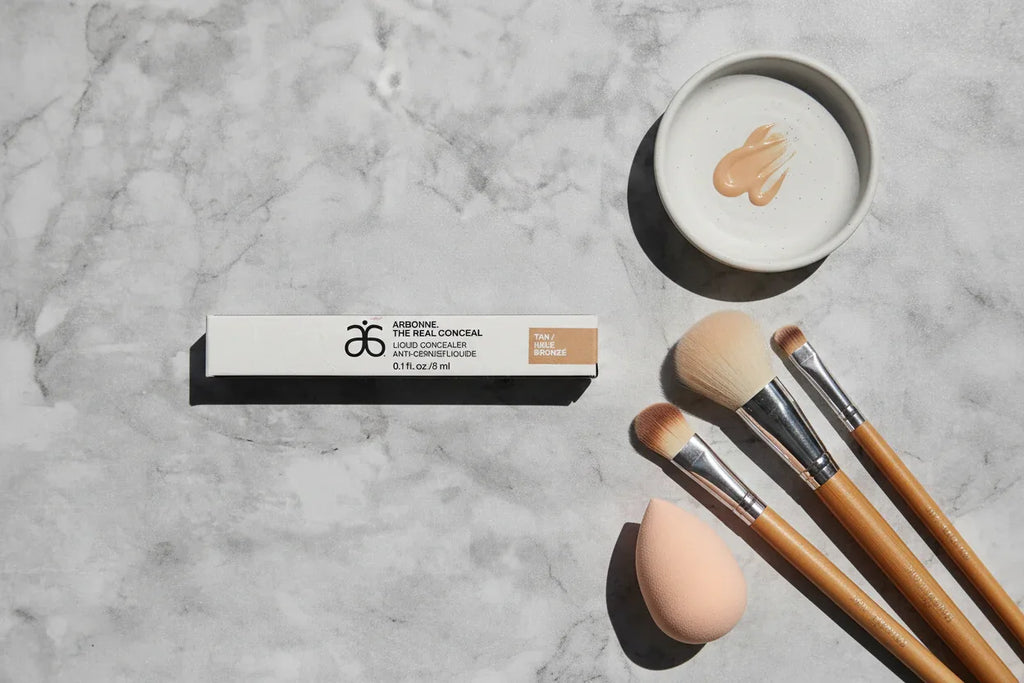 Arbonne The Real Conceal Liquid Concealer Tan flat lay on slate surface with blending brushes