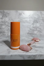 Modern minimalist vanity layout showing small orange brightening serum bottle next to rose quartz facial roller on white marble