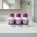 Three MiraLAX Laxative Powder bottles (4.1 oz each) factory shrink-wrapped together — white bottles with purple caps and dark purple MiraLAX labels — standing upright on a clean white bathroom countertop with soft natural lighting and blurred medicine cabinet