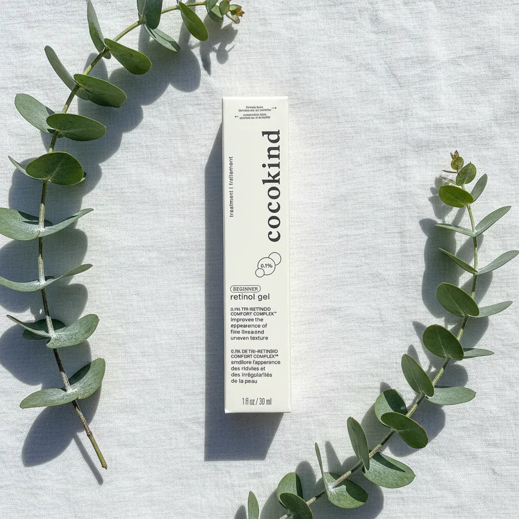 Overhead flat lay of Cocokind Beginner Retinol Gel white box on white linen with fresh green eucalyptus sprigs, bright natural light