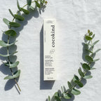 Overhead flat lay of Cocokind Beginner Retinol Gel white box on white linen with fresh green eucalyptus sprigs, bright natural light