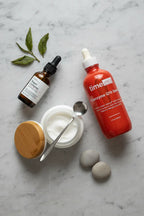 Professional flat-lay composition of premium skincare essentials including a dark amber glass serum bottle on smooth grey stone surface