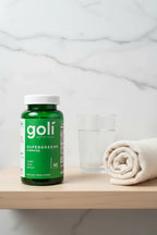 Goli Supergreens Gummies jar on minimalist bathroom vanity shelf with rolled cotton towel and glass of water — More4less Outlet