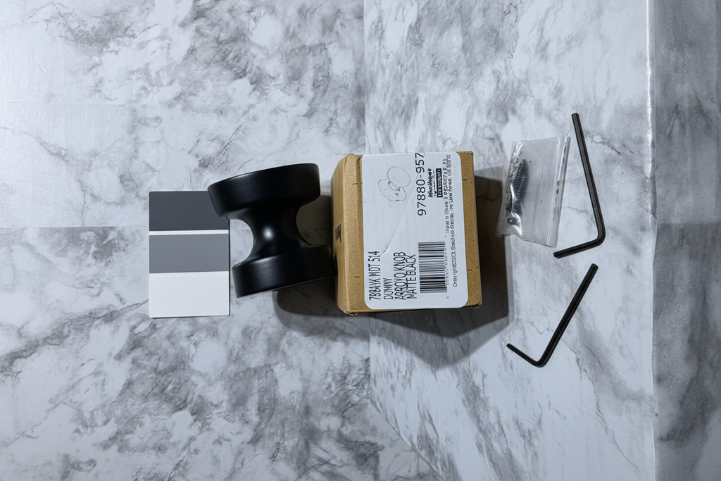 Kwikset Arroyo matte black dummy knob flatlay on white marble with retail box hex wrench and charcoal paint swatches - More4less Outlet