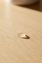 Detail shot of a clear liquid moisture drop being absorbed into smooth light wood surface in warm natural lighting