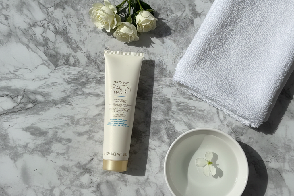 Slim white hand cream tube next to soft white towel, bowl of water, and fresh flowers on marble