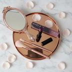 Mary Kay Lip Suede Lipstick Mulberry Muse in black gloss tube on a rose-gold tray with makeup brushes, a small mirror, and pink rose petals on a white marble surface — feminine beauty flat-lay