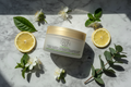 Frosted white luxury body cream jar next to white tea flowers and lemon slices on marble