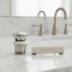 American Standard satin nickel drain assembly and overflow cover plate laid out side by side on a white marble bathroom countertop