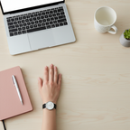 Minimalist desk flat-lay with woman's hand wearing black strap watch next to pink notebook and laptop