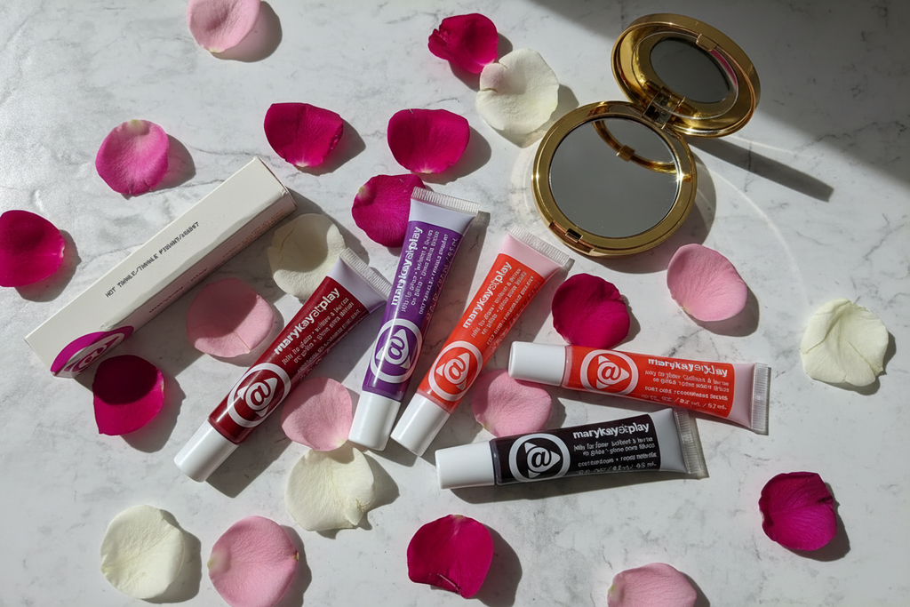 Colorful lip gloss tubes in jewel-toned shades scattered on white marble with rose petals and compact mirror – editorial beauty photography