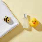 Lifestyle — soft white nursery flatlay with cologne bottle beside yellow rubber duck, white baby towel, and bee plush on pastel yellow background — editorial for visual context only