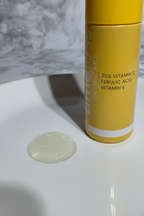 A drop of clear Vitamin C serum on smooth white porcelain surface next to a bright yellow skincare bottle in soft studio lighting