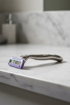 Sleek platinum metal women's razor resting on a clean white marble bathroom counter with soft dramatic lighting
