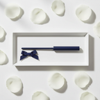 Lifestyle image — navy blue perfume wand on white gift tray beside navy ribbon bow and white rose petals, elegant gifting flatlay for visual context only