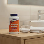 NOW Foods Inositol Powder 8oz bottle on light-oak bathroom vanity shelf with aesthetic glass of water and white cotton hand towel