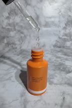 Glass pipette with clear serum droplets held above a small orange skincare bottle on white marble in soft clean lighting